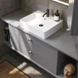 Badezimmer mit schrägen Wänden, grauem Waschtisch und Oberschrank, maßgefertigte Arbeitsplatten, modernes quadratisches Aufsatzwaschbecken, quadratischer Spiegel mit Beleuchtung, rohe Betonwände und -böden verleihen einen urbanen Ausdruck