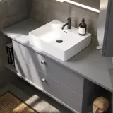 Badezimmer mit schrägen Wänden, grauem Waschtisch und Oberschrank, maßgefertigte Arbeitsplatten, modernes quadratisches Aufsatzwaschbecken, quadratischer Spiegel mit Beleuchtung, rohe Betonwände und -böden verleihen einen urbanen Ausdruck
