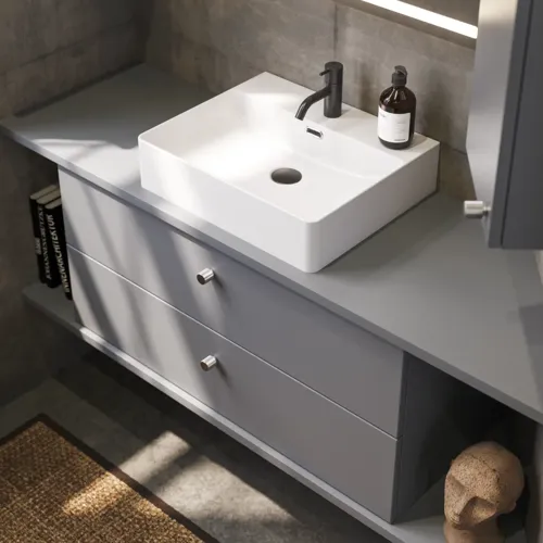 Badezimmer mit schrägen Wänden, grauem Waschtisch und Oberschrank, maßgefertigte Arbeitsplatten, modernes quadratisches Aufsatzwaschbecken, quadratischer Spiegel mit Beleuchtung, rohe Betonwände und -böden verleihen einen urbanen Ausdruck