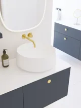 Elegante und moderne Badezimmer Inspiration mit schrägem Dach, wandmontierten blauen Waschtischunterschränken und Unterschränken, rundem Waschbecken mit Messingarmatur, beleuchtetem Spiegel und natürlichem Licht