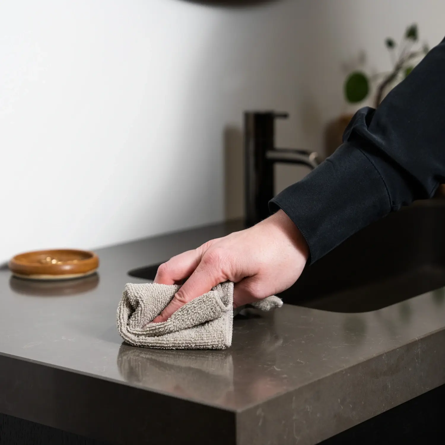 Reinigung und Pflege von Badezimmermöbeln: Eine Person wischt vorsichtig eine dunkle Badezimmer-Arbeitsplatte mit einem beigefarbenen Mikrofasertuch ab, um Schmutz und Wasserspritzer zu entfernen.